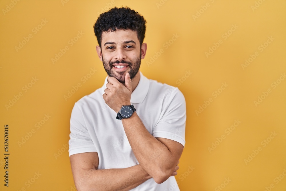 Arab man standing over yellow background looking confident at the camera smiling with crossed arms and hand raised on chin. thinking positive. Arab man standing over yellow background looking confident at the camera smiling with crossed arms and hand raised on chin. thinking positive.