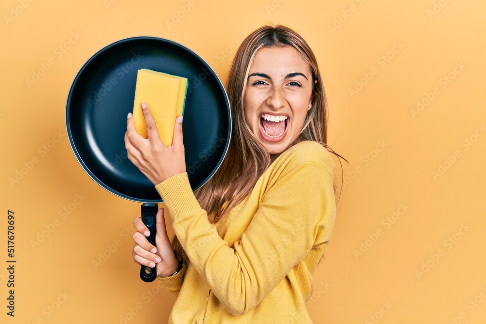 Beautiful hispanic woman cleaning cooking pan with scourer celebrating crazy and amazed for success with open eyes screaming excited. Beautiful hispanic woman cleaning cooking pan with scourer celebrating crazy and amazed for success with open eyes screaming excited.