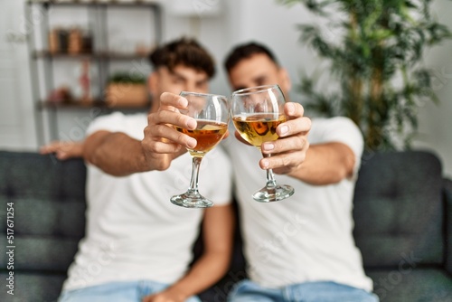 Two hispanic men couple toasting with glass of wine sitting on sofa at home