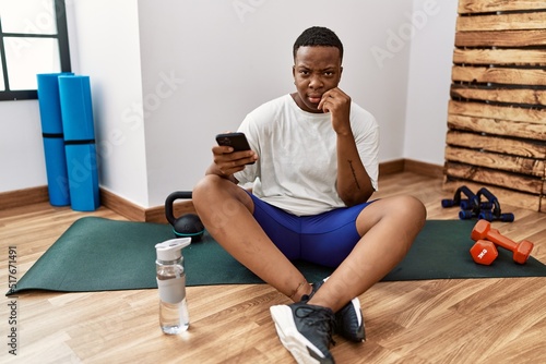 Young african man sitting on training mat at the gym using smartphone looking stressed and nervous with hands on mouth biting nails. anxiety problem.