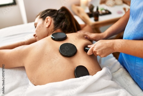 Two women therapist and patient having back massage session using black stones at beauty center