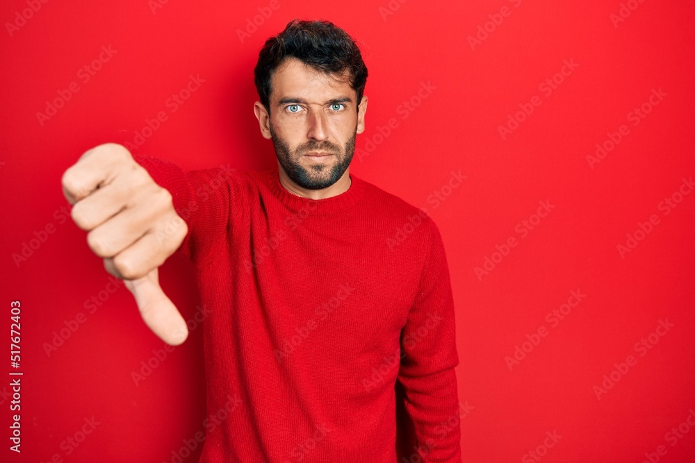 Handsome man with beard wearing casual red sweater looking unhappy and angry showing rejection and negative with thumbs down gesture. bad expression. Handsome man with beard wearing casual red sweater looking unhappy and angry showing rejection and negative with thumbs down gesture. bad expression.