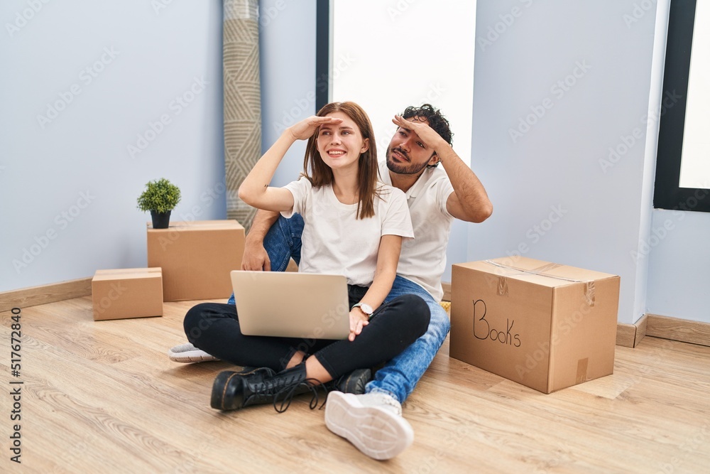 Young couple using laptop at new home very happy and smiling looking far away with hand over head. searching concept. Young couple using laptop at new home very happy and smiling looking far away with hand over head. searching concept.