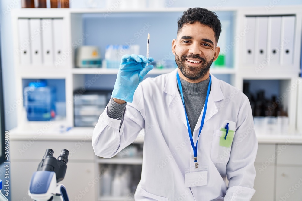 Hispanic man with beard working at scientist laboratory holding syringe looking positive and happy standing and smiling with a confident smile showing teeth Hispanic man with beard working at scientist laboratory holding syringe looking positive and happy standing and smiling with a confident smile showing teeth