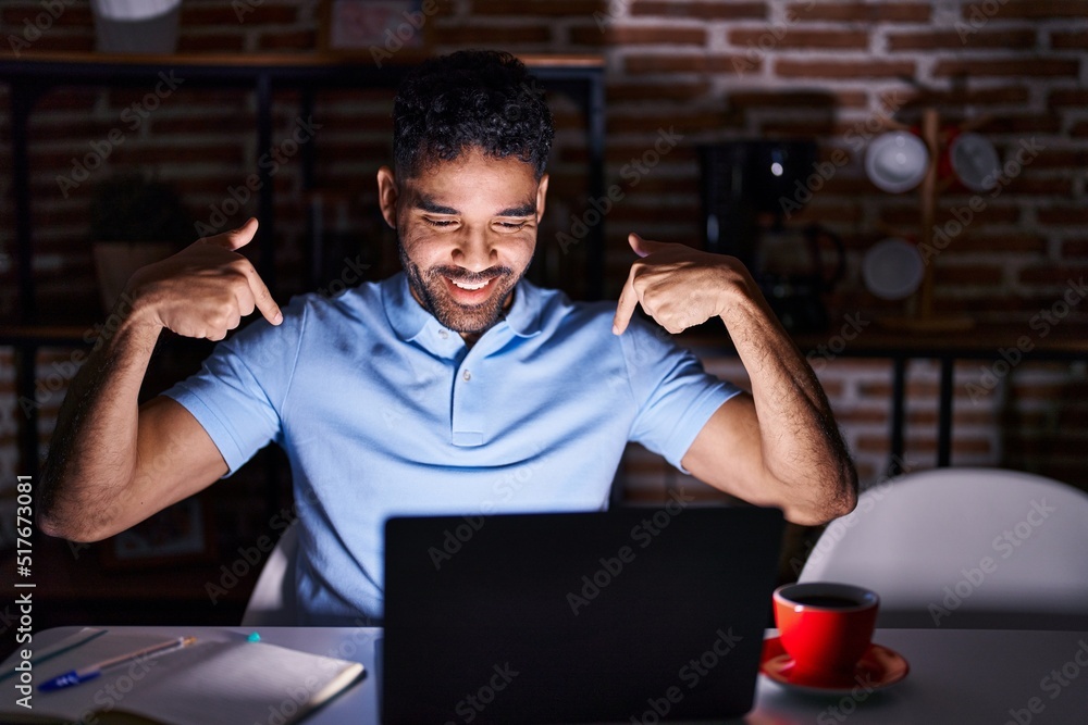 Hispanic man with beard using laptop at night looking confident with smile on face, pointing oneself with fingers proud and happy.