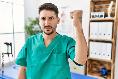 Young physiotherapist man working at pain recovery clinic angry and mad raising fist frustrated and furious while shouting with anger. rage and aggressive concept.