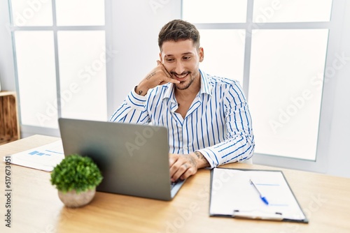 Young handsome man with beard working at the office using computer laptop smiling doing phone gesture with hand and fingers like talking on the telephone. communicating concepts.