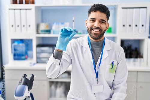 Hispanic man with beard working at scientist laboratory holding syringe looking positive and happy standing and smiling with a confident smile showing teeth