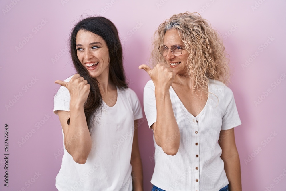 Mother and daughter standing together over pink background smiling with happy face looking and pointing to the side with thumb up. Mother and daughter standing together over pink background smiling with happy face looking and pointing to the side with thumb up.