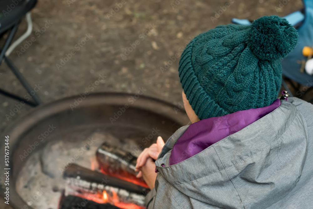 Camping lifestyle concept. Girl wearing beanie hat wariming hands over the campfire on a camping site
