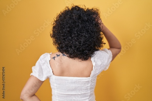Young brunette woman with curly hair standing over yellow background backwards thinking about doubt with hand on head