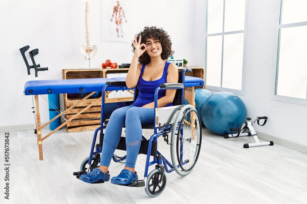 Obraz premium Young middle eastern woman sitting on wheelchair at physiotherapy clinic smiling positive doing ok sign with hand and fingers. successful expression.