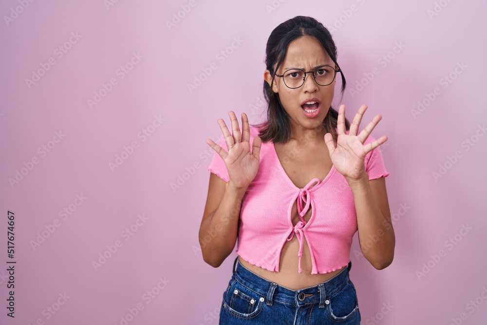 Hispanic young woman standing over pink background wearing glasses afraid and terrified with fear expression stop gesture with hands, shouting in shock. panic concept. Hispanic young woman standing over pink background wearing glasses afraid and terrified with fear expression stop gesture with hands, shouting in shock. panic concept.