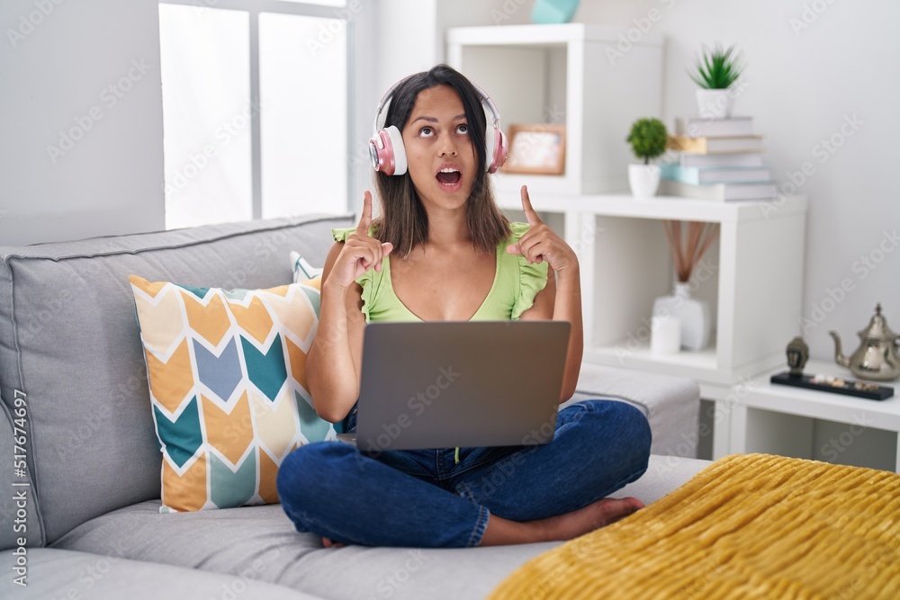 Hispanic young woman using laptop at home amazed and surprised looking up and pointing with fingers and raised arms. Hispanic young woman using laptop at home amazed and surprised looking up and pointing with fingers and raised arms.