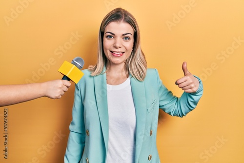 Beautiful caucasian woman being interviewed by reporter holding microphone smiling happy and positive  thumb up doing excellent and approval sign
