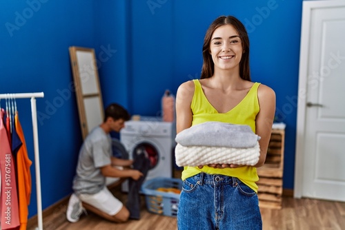 Fototapeta Naklejka Na Ścianę i Meble -  Young brunette woman holding folded laundry looking positive and happy standing and smiling with a confident smile showing teeth