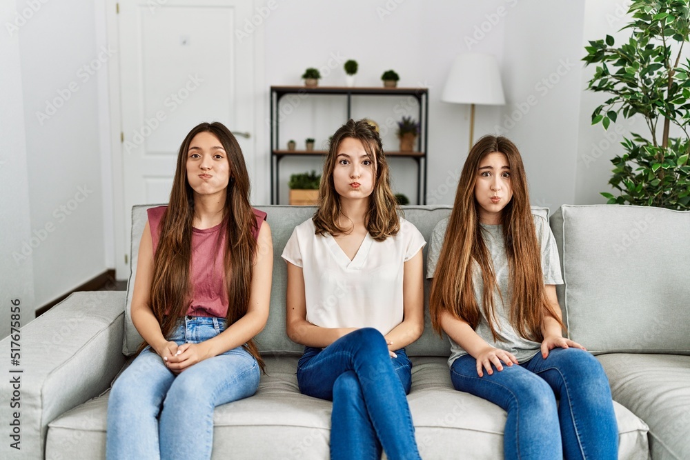 Group of three hispanic girls sitting on the sofa at home puffing cheeks with funny face. mouth inflated with air, crazy expression. Group of three hispanic girls sitting on the sofa at home puffing cheeks with funny face. mouth inflated with air, crazy expression.
