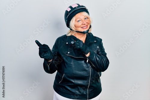 Middle age blonde woman holding motorcycle helmet smiling and looking at the camera pointing with two hands and fingers to the side.
