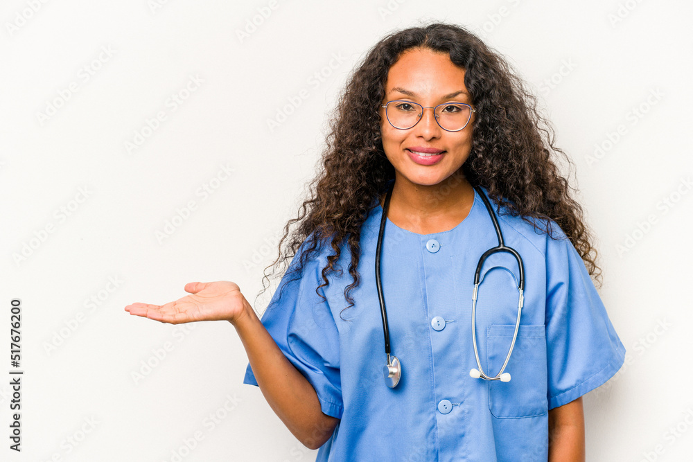 Young hispanic nurse woman isolated on white background showing a copy space on a palm and holding another hand on waist. Young hispanic nurse woman isolated on white background showing a copy space on a palm and holding another hand on waist.