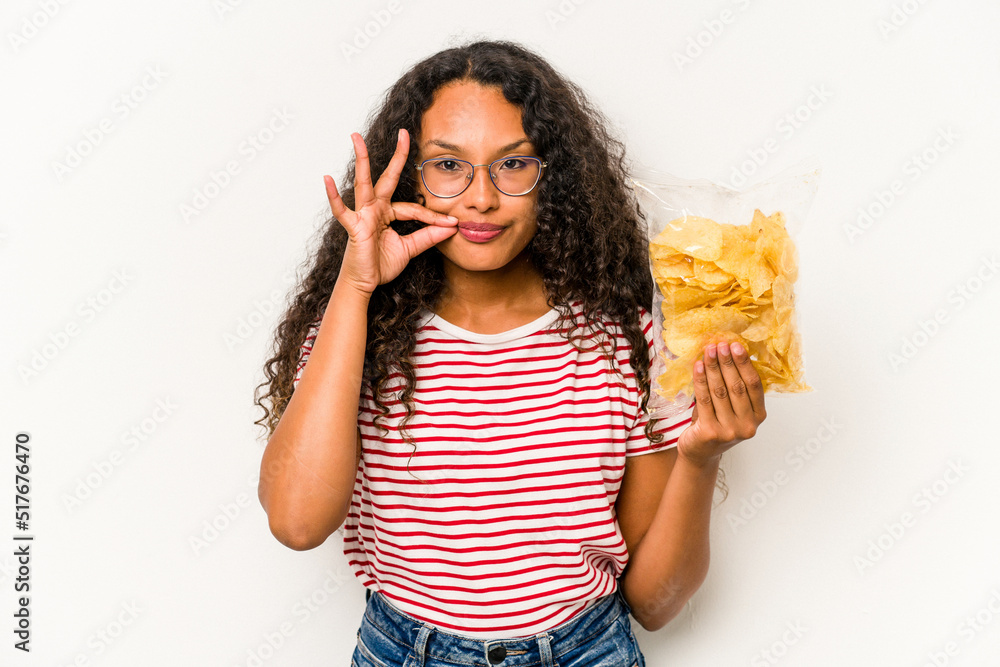 Young hispanic woman holding crips isolated on white background with fingers on lips keeping a secret. Young hispanic woman holding crips isolated on white background with fingers on lips keeping a secret.
