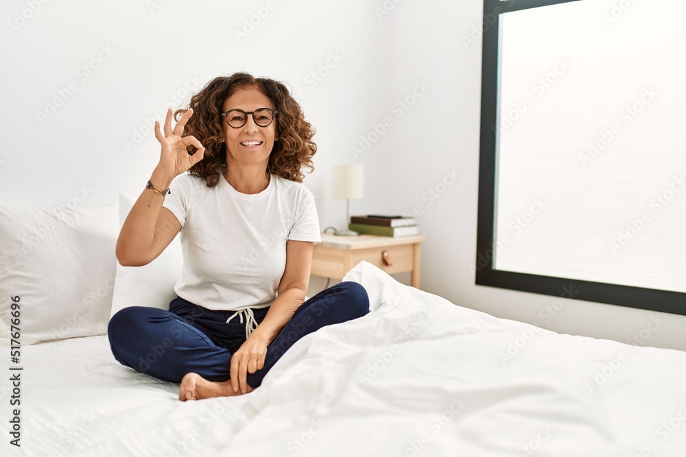 Middle age hispanic woman sitting on the bed at home smiling positive doing ok sign with hand and fingers. successful expression. Middle age hispanic woman sitting on the bed at home smiling positive doing ok sign with hand and fingers. successful expression.