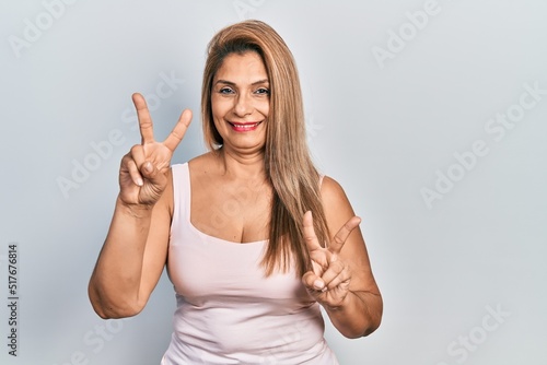 Middle age hispanic woman wearing casual style with sleeveless shirt smiling looking to the camera showing fingers doing victory sign. number two.