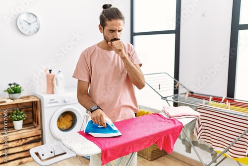 Young hispanic man ironing clothes at home feeling unwell and coughing as symptom for cold or bronchitis. health care concept.