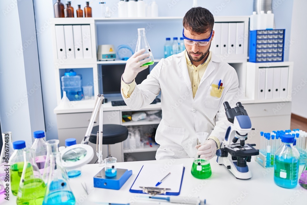 Young hispanic man wearing scientist uniform measuring liquid at laboratory Young hispanic man wearing scientist uniform measuring liquid at laboratory