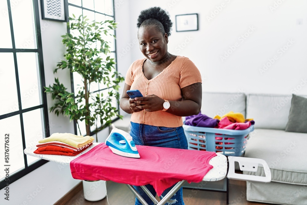 Young african american woman using smartphone ironing clothes at home Young african american woman using smartphone ironing clothes at home