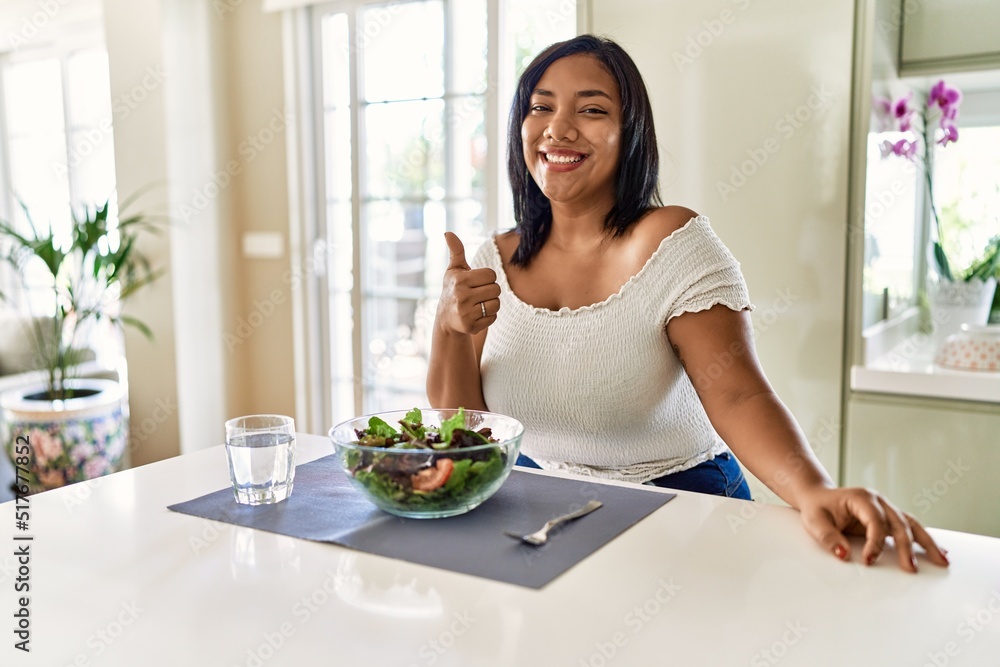 Young hispanic woman eating healthy salad at home doing happy thumbs up gesture with hand. approving expression looking at the camera showing success. Young hispanic woman eating healthy salad at home doing happy thumbs up gesture with hand. approving expression looking at the camera showing success.