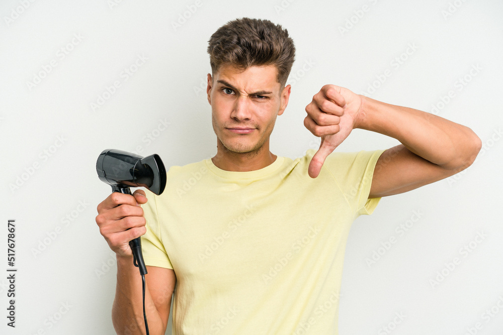 Young caucasian man holding hairdryer isolated on white background showing a dislike gesture, thumbs down. Disagreement concept. Young caucasian man holding hairdryer isolated on white background showing a dislike gesture, thumbs down. Disagreement concept.