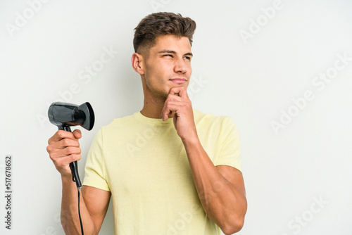 Young caucasian man holding hairdryer isolated on white background looking sideways with doubtful and skeptical expression.