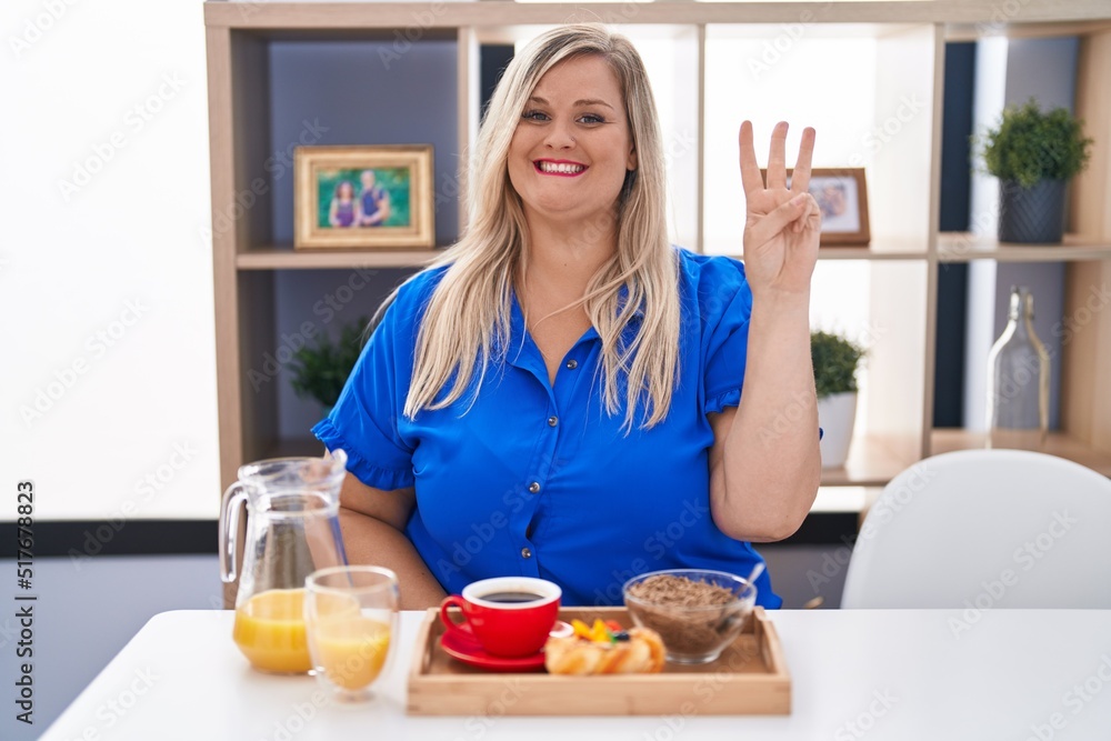Caucasian plus size woman eating breakfast at home showing and pointing up with fingers number three while smiling confident and happy.