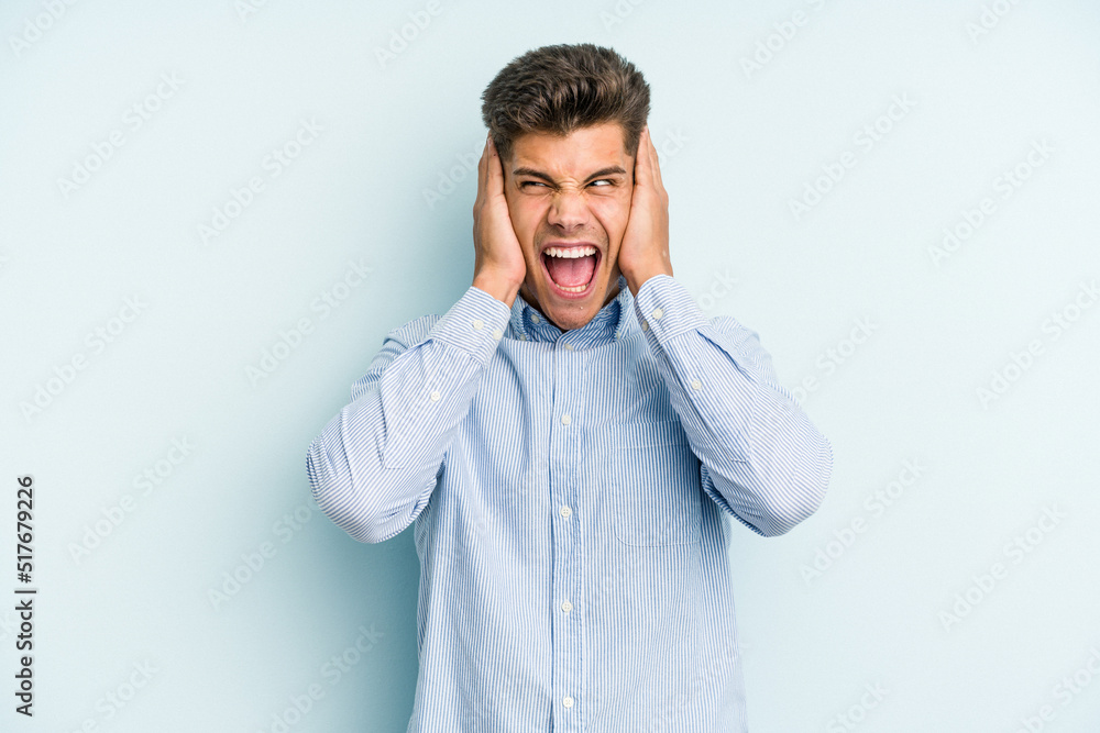 Young caucasian man isolated on blue background covering ears with hands trying not to hear too loud sound. Young caucasian man isolated on blue background covering ears with hands trying not to hear too loud sound.