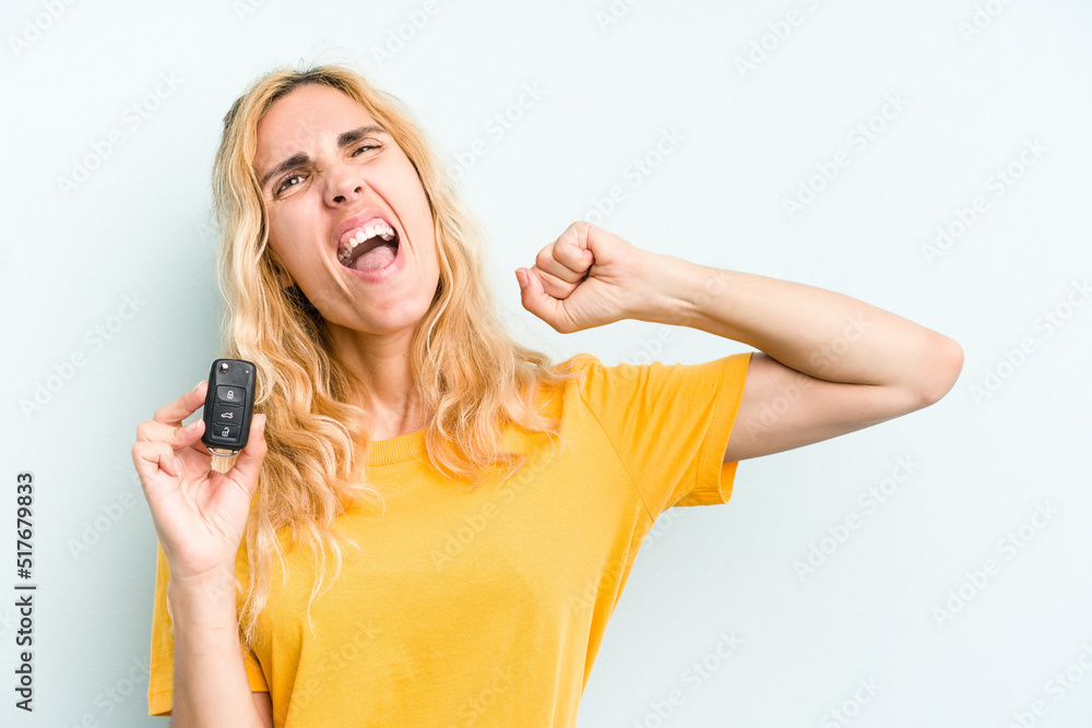 Young caucasian woman holding car keys isolated on blue background raising fist after a victory, winner concept. Young caucasian woman holding car keys isolated on blue background raising fist after a victory, winner concept.