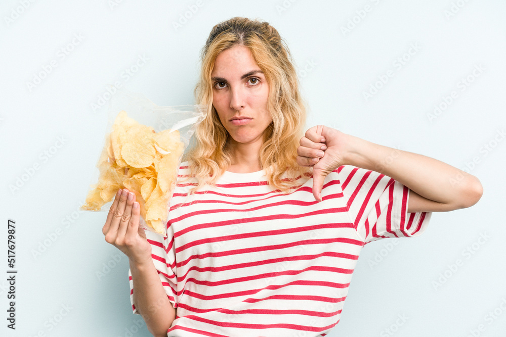 Young caucasian woman holding a bag of chips isolated on blue background showing a dislike gesture, thumbs down. Disagreement concept. Young caucasian woman holding a bag of chips isolated on blue background showing a dislike gesture, thumbs down. Disagreement concept.