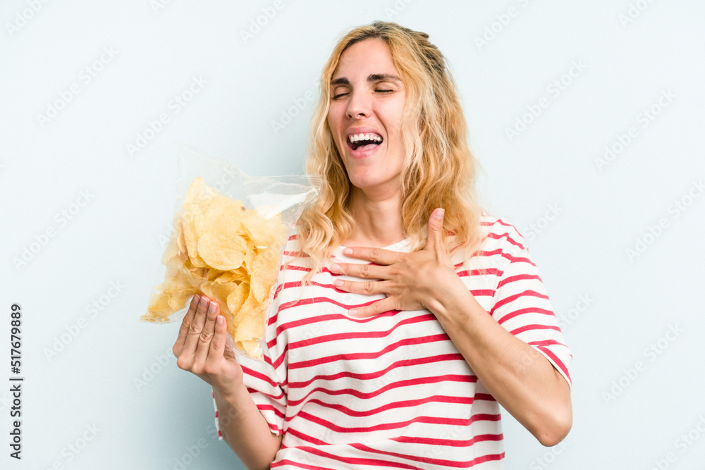 Young caucasian woman holding a bag of chips isolated on blue background laughs out loudly keeping hand on chest. Young caucasian woman holding a bag of chips isolated on blue background laughs out loudly keeping hand on chest.