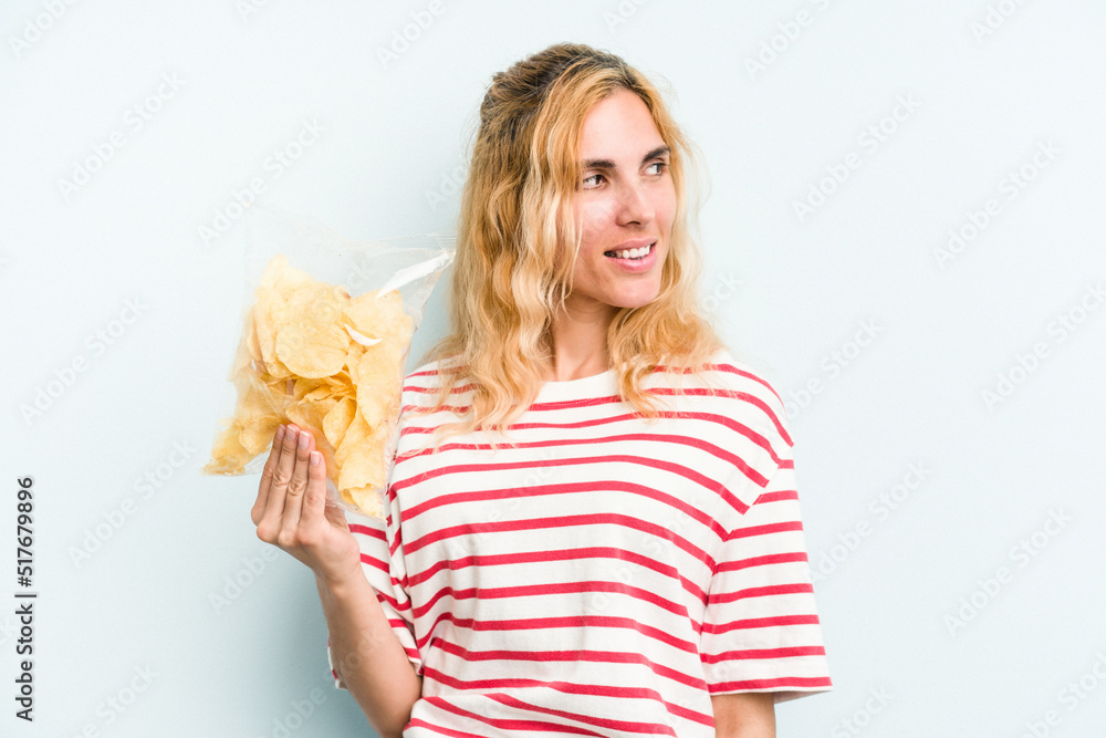 Young caucasian woman holding a bag of chips isolated on blue background looks aside smiling, cheerful and pleasant. Young caucasian woman holding a bag of chips isolated on blue background looks aside smiling, cheerful and pleasant.