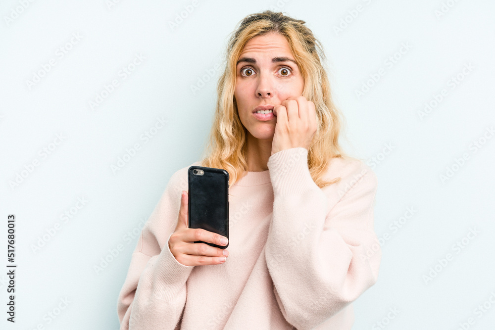 Young caucasian woman holding mobile phone isolated on blue background biting fingernails, nervous and very anxious. Young caucasian woman holding mobile phone isolated on blue background biting fingernails, nervous and very anxious.