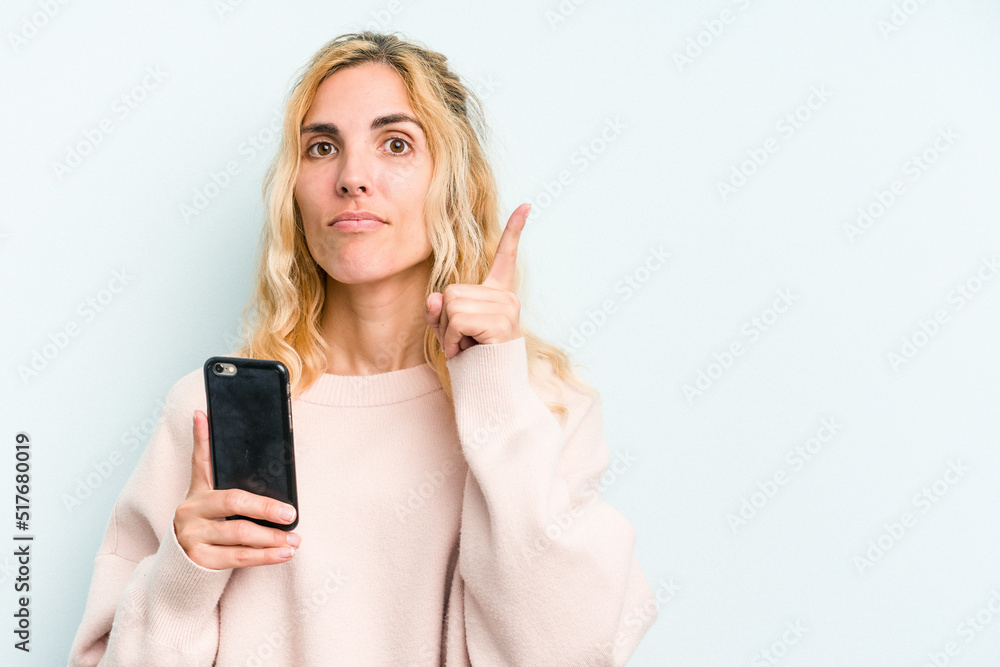 Young caucasian woman holding mobile phone isolated on blue background showing number one with finger. Young caucasian woman holding mobile phone isolated on blue background showing number one with finger.