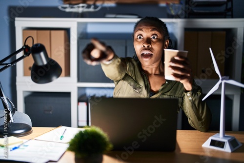African woman working using computer laptop at night pointing with finger surprised ahead, open mouth amazed expression, something on the front