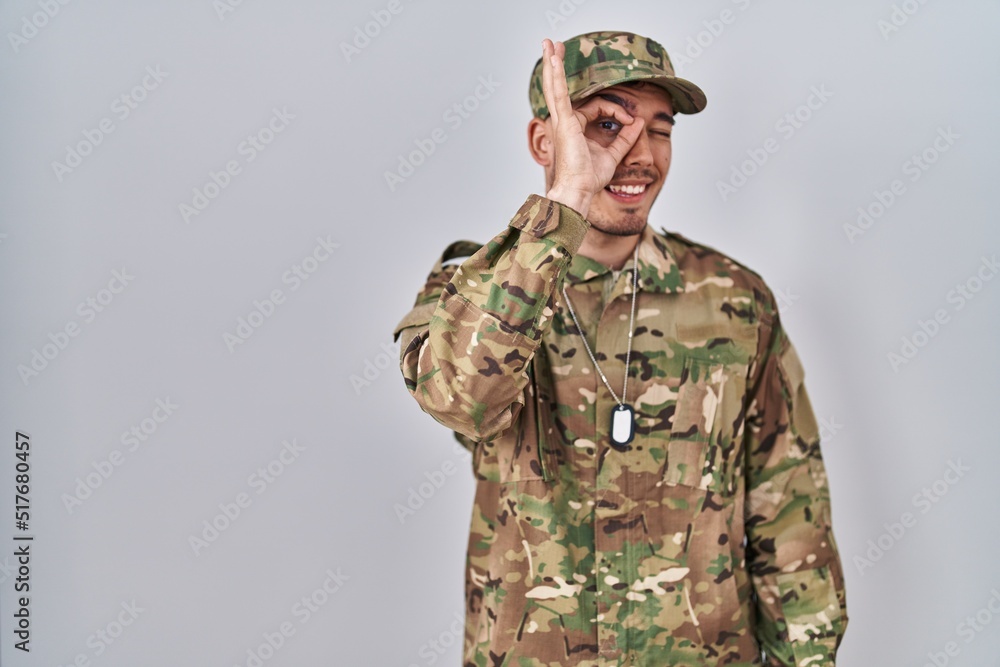 Young hispanic man wearing camouflage army uniform doing ok gesture with hand smiling, eye looking through fingers with happy face.