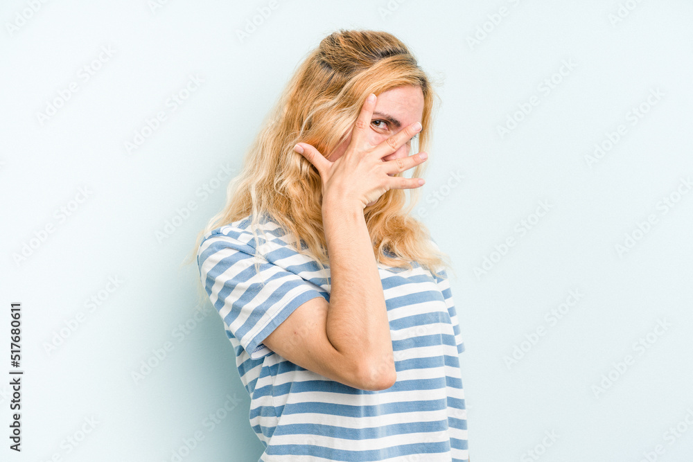 Young caucasian woman isolated on blue background blink at the camera through fingers, embarrassed covering face. Young caucasian woman isolated on blue background blink at the camera through fingers, embarrassed covering face.