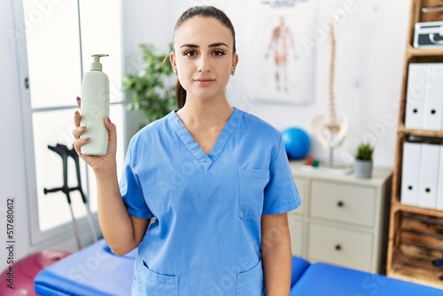 Young physiotherapist woman holding massage body lotion thinking attitude and sober expression looking self confident