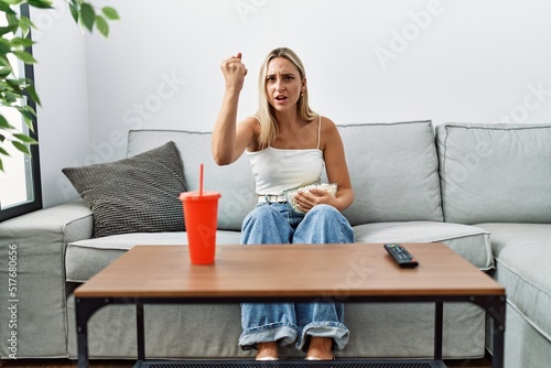 Young blonde woman eating popcorn sitting on the sofa annoyed and frustrated shouting with anger, yelling crazy with anger and hand raised