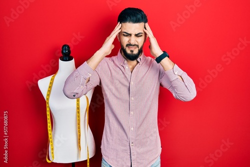 Handsome tailor man with beard standing by manikin with hand on head, headache because stress. suffering migraine.