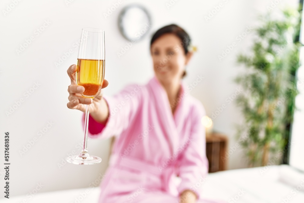 Middle age hispanic woman toasting with champagne sitting on massage table at beauty center Middle age hispanic woman toasting with champagne sitting on massage table at beauty center