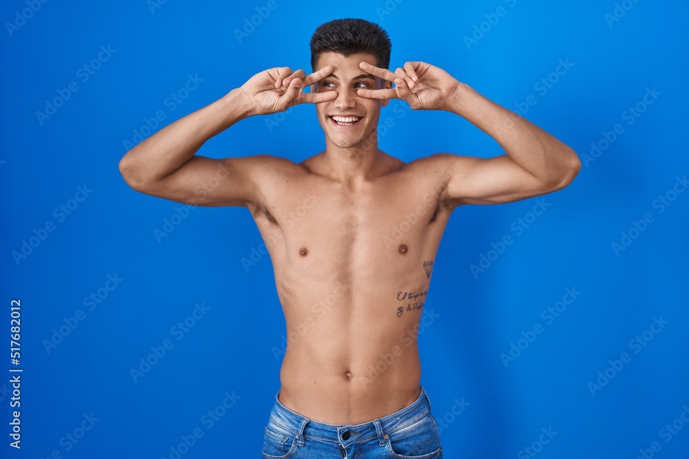 Young hispanic man standing shirtless over blue background doing peace symbol with fingers over face, smiling cheerful showing victory