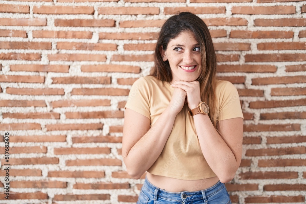 Young brunette woman standing over bricks wall laughing nervous and excited with hands on chin looking to the side Young brunette woman standing over bricks wall laughing nervous and excited with hands on chin looking to the side