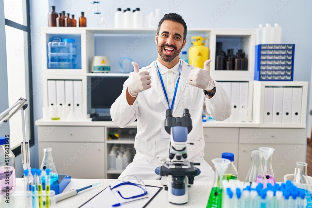 Young hispanic man with beard working at scientist laboratory success sign doing positive gesture with hand, thumbs up smiling and happy. cheerful expression and winner gesture. Young hispanic man with beard working at scientist laboratory success sign doing positive gesture with hand, thumbs up smiling and happy. cheerful expression and winner gesture.